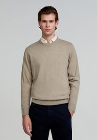 Beige gebreide trui met een ronde halslijn en lange mouwen, gecombineerd met een lichtgekleurde overhemden. Soepele textuur en relaxed pasvorm.