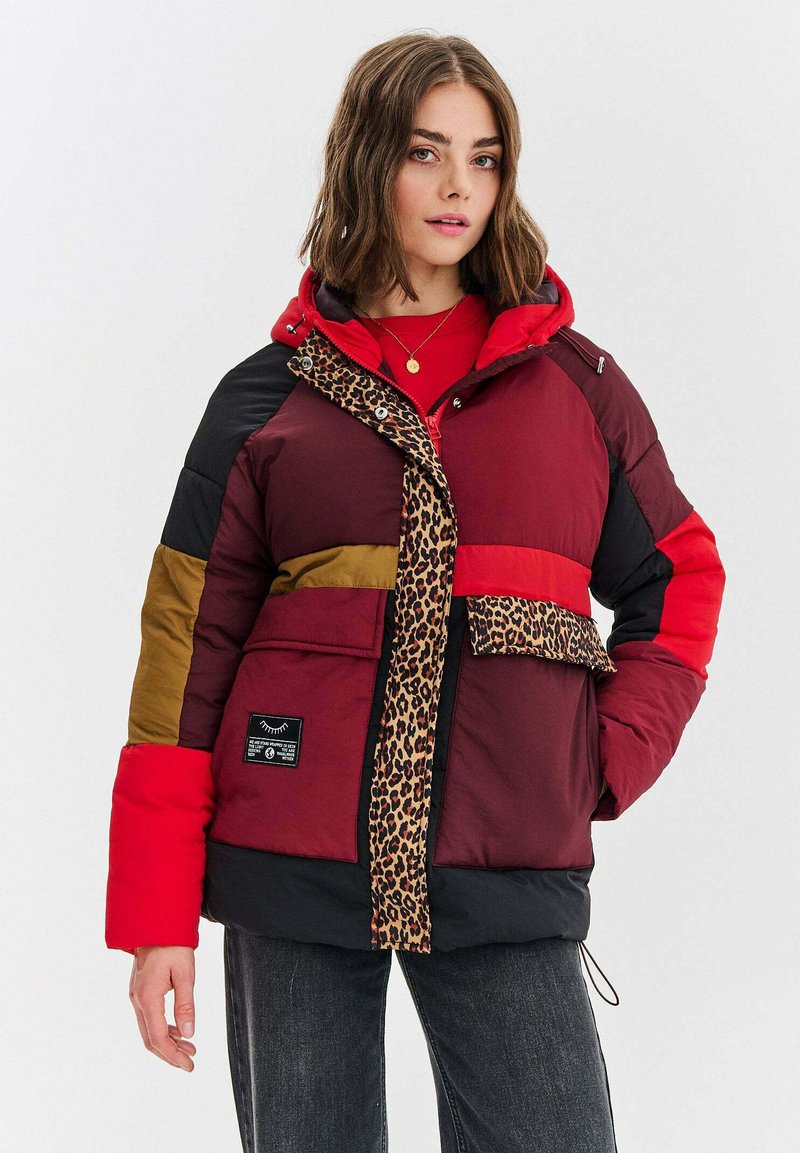 Žena oblečená v mnohobarevnej prešívanej bunde s červenými, baklažánovými, čiernymi a olivovými časťami, doplnená o leopardí potlač. K tomu má na sebe tmavé džínsy.