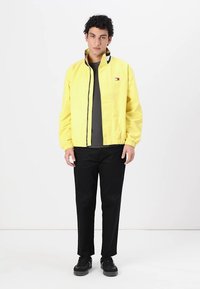 Gelbe leichte Jacke mit Stehkragen, schwarzer Einfassung und elastischen Bündchen, kombiniert mit schwarzen Hosen und dunklen Sneakers.