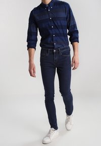 Námořnická košile na knoflíky s horizontálními pruhy, zkombinovaná s tmavými džíny a bílými teniskami. Oblečení má klasický límec a přiléhavý střih.