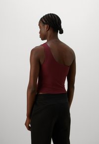 Vrouw met gevlochten haar, gekleed in een bordeauxrode één-schouder tanktop en zwarte broek, met de rug naar de camera gekeerd tegen een effen lichte achtergrond.