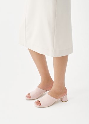 Jambes portant une jupe midi couleur crème et des mules à talon large ouvertes de couleur rose pâle sur fond blanc.