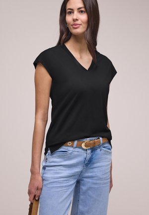 Femme portant un haut sans manches noir à col en V, un jean bleu clair avec une ceinture marron, tenant un sac à main marron sur un fond neutre.