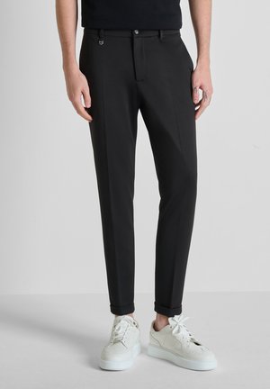 Hombre con pantalones negros ajustados con puños y zapatillas blancas, de pie frente a un fondo claro y sencillo.