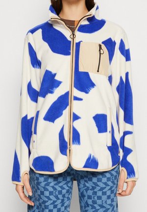 Bluza polarowa z białą podstawą i niebieskimi abstrakcyjnymi łatami, z beżowym zamkiem błyskawicznym i kieszenią na piersi, noszona z niebieskimi spodniami w wzory.