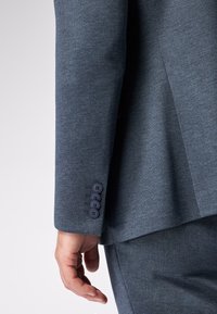 Giacca blu su misura con una texture liscia, dotata di quattro bottoni navy sul polsino e un design strutturato, che mostra la manica e la mano.