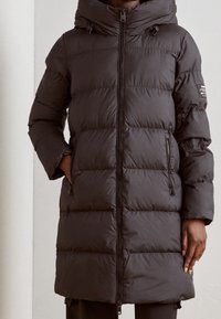 Černá péřová bunda se zapínáním na zip, kapucí a bočními kapsami. Obsahuje prošívaný design s rovnoměrně rozloženými panely a elastickými manžetami.