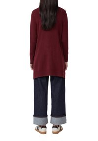 Femme avec de longs cheveux noirs portant un long pull bourgogne, un large jean bleu marine retroussé et des baskets blanches, se tenant de dos.