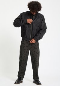 Giubbotto bomber nero con zip frontale, polsini a coste e un emblema ricamato. Abbinato a pantaloni oliva con stampa di pipistrelli. Scarpe nere.