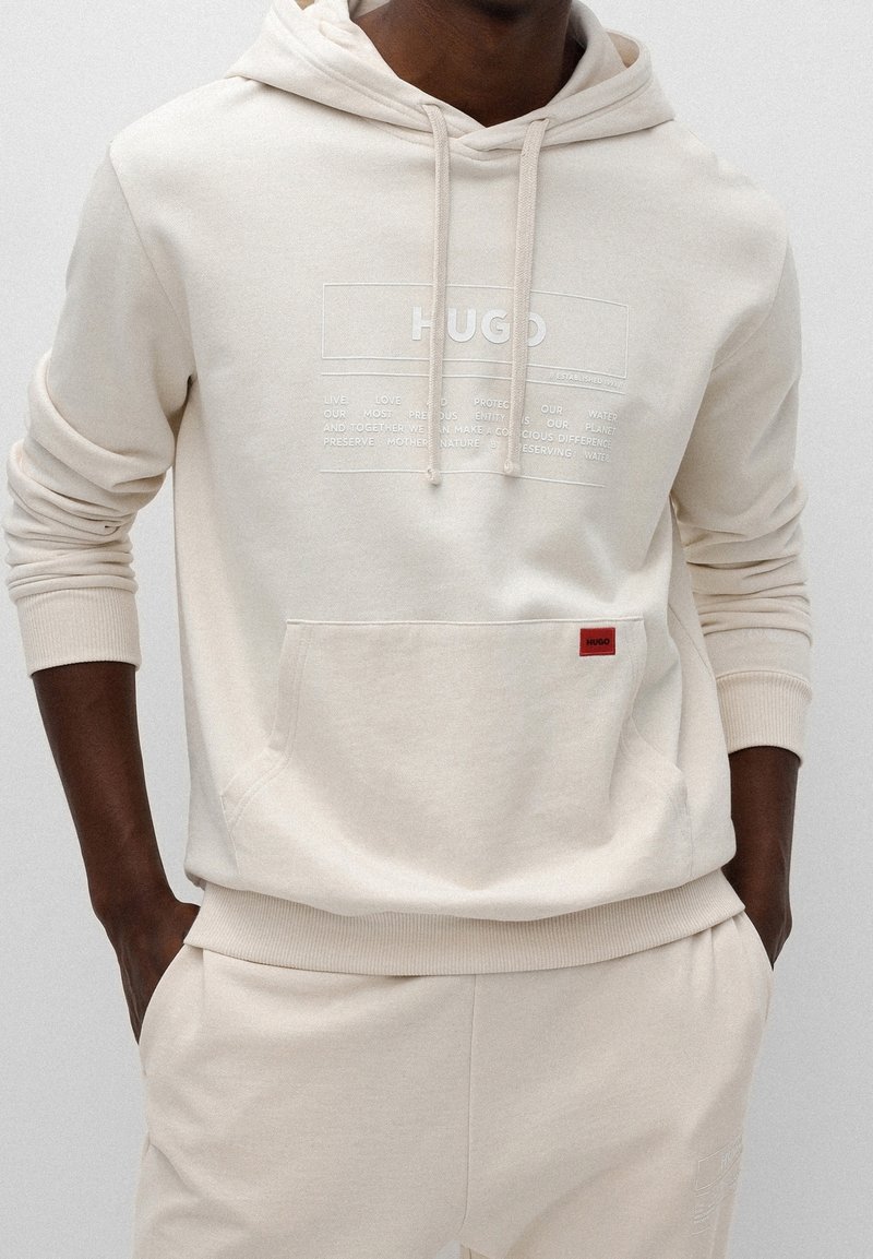 Felpa beige con tasca frontale, cordini e stampa del logo bianca. Polsini e orlo a coste, tessuto morbido, abbinata a pantaloni beige coordinati.