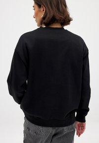 Sudadera negra de cuello redondo con hombros caídos, puños y dobladillo de canalé, textura suave y un corte relajado, combinada con jeans grises.