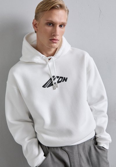 Sudadera blanca con un gran gráfico negro de "ICON" en el frente, cordones con puntas de metal y puños acanalados. Llevada sobre pantalones grises.