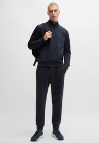 Navyblå atletisk träningsdräkt i slät tyg, med dragkedja fram, ribbade ärmslut och matchande sneakers. Rygga på modellen.
