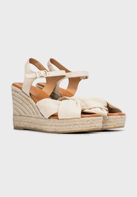 Sandalias de cuña beige con un empeine de tela anudada, correa de tobillo ajustable y plataforma de yute trenzado. Plantilla de cuero suave para mayor comodidad.