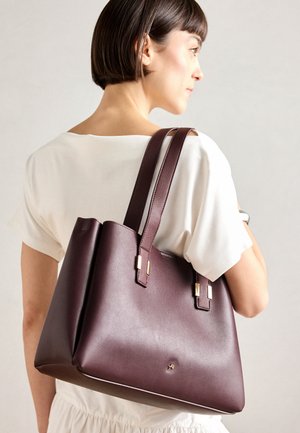 Burgundy leather tote bag with two handles, gold hardware accents, structured shape, and smooth texture. Minimalist design with a logo.