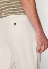 Ljusbeige byxor med fiskbensmönster, med en bakficka med en brun knappdetalj. Texturerat tyg med en skräddarsydd passform.