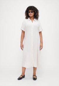 White, knee-length shirt dress made of lightweight fabric; features short rolled sleeves, a button front, and a relaxed fit. Black flats worn.