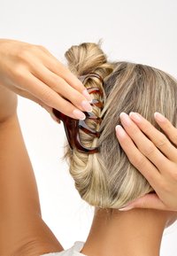Fibbia per capelli tartarugata che tiene in posizione un chignon attorcigliato. Finitura liscia e lucida con più denti per la presa. Mani posizionate su entrambi i lati dei capelli.