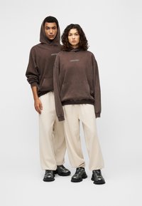 Δύο μοντέλα φορούν φαρδιά καφέ φούτερ με κουκούλα και μπροστινές τσέπες, σε συνδυασμό με εκρού παντελόνες φαρδιές. Φορουν μαύρα chunky αθλητικά παπούτσια.