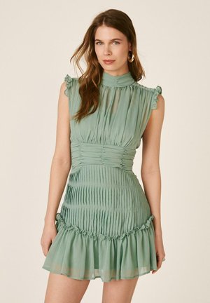 Mujer con cabello largo y castaño que lleva un vestido sin mangas, de cuello alto, verde salvia plisado con dobladillo con volantes sobre un fondo beige liso.