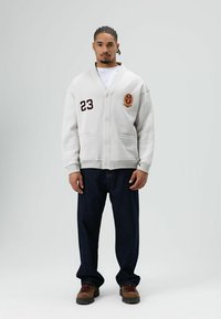 Lichtgrijze cardigan met knoopsluiting, geborduurd nummer "23" en een wapen. Gecombineerd met donkerblauwe jeans en bruine laarzen. Soepele textuur.