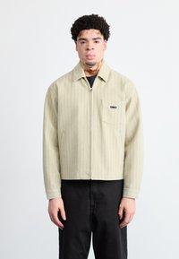 Jachetă bej cu dungi, cu fermoar în față și un buzunar pe partea stângă a pieptului, având un logo negru. Asortată cu pantaloni negri.