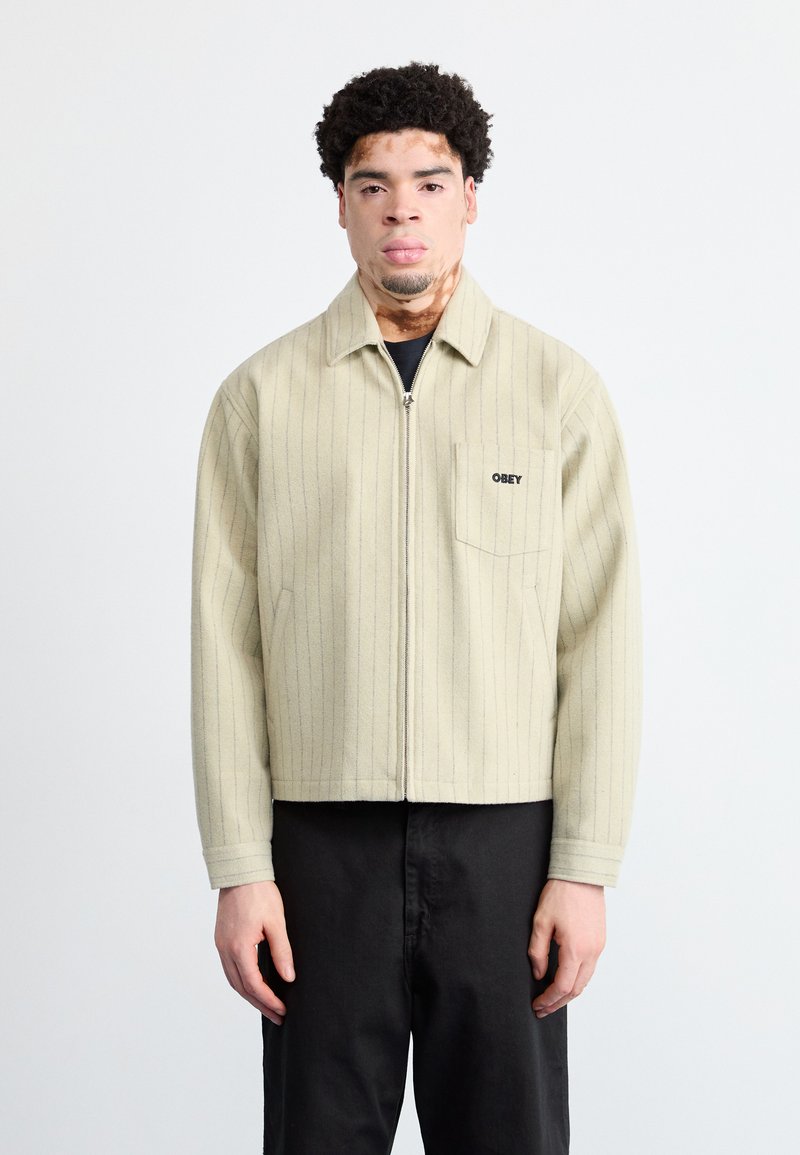 Jachetă bej cu dungi, cu fermoar în față și un buzunar pe partea stângă a pieptului, având un logo negru. Asortată cu pantaloni negri.