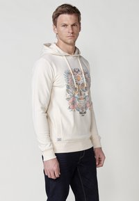 Béživa dukserica s kapuljačom, s grafičkim otiskom lubanje, cvjetnim detaljima i natpisom "TROPIC VIBES" u kontrastnoj boji. Mekani materijal, kengurska džep.