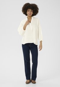 Blusa bianca in lino con placca a metà bottone e colletto arricciato; abbinata a jeans blu scuro e ballerine con stampa leopardata.