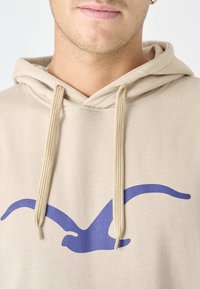 Felpa beige con cappuccio e lacci, caratterizzata da una grande grafica di un uccello blu sul davanti. Il tessuto sembra morbido e ha una texture liscia.