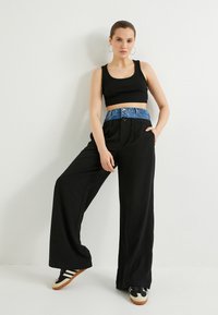 Pantaloni neri a gamba larga con dettaglio in denim in vita, abbinati a un top corto nero e sneakers nere con suola crema e marrone.