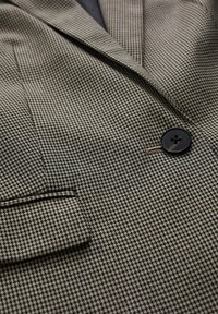 Gros plan d'un blazer marron et noir à motif pied-de-poule avec bouton noir et rabat de poche bordé.