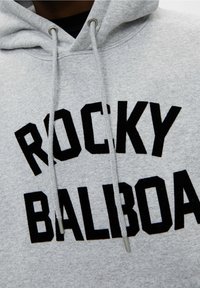 Sweat à capuche gris avec texte noir en gras "ROCKY BALBOA" sur la poitrine et cordons gris avec embouts en métal.