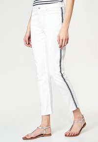 Bílé džíny s krátkým střihem, zdobené černými bočními pruhy, pěti kapsami a knoflíkovým zapínáním. V kombinaci se stříbrnými plochými sandály.