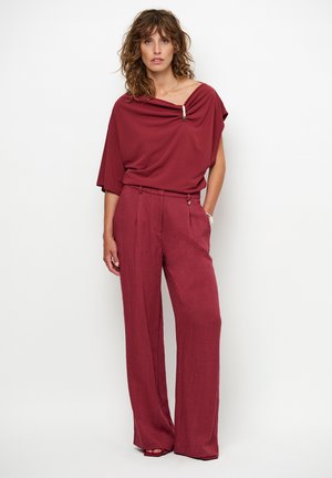 Bordeauxrotes Oberteil mit schulterfreiem Design und einer dekorativen Brosche; kombiniert mit hoch taillierten, weit geschnittenen Hosen in demselben Bordeauxfarbton.