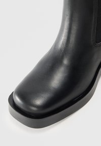 Botín negro de cuero hasta el tobillo con punta redonda, paneles laterales elásticos y una gruesa suela de goma negra. Textura suave con un acabado elegante.