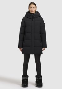Fekete párnázott parka, kapucnival, cipzárral, oldalsó zsebekkel és texturált felülettel. Fekete leggingsszel és csizmával viselve.