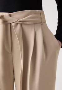 Pantalon beige taille haute avec plis et ceinture en tissu nouée, porté avec un haut noir à manches longues et une main dans la poche.