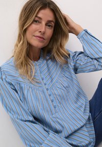 Blusa azzurro chiaro con strisce verticali bianche e dorate, design con bottoni frontali, dettagli arricciati e maniche lunghe. Tessuto morbido.