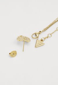 Il set di gioielli in metallo dorato include un orecchino a perno con strass trasparenti e una catena con un ciondolo triangolare che presenta il nome del marchio.