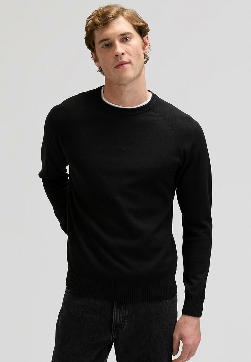 Maglione nero lavorato a maglia con collo rotondo, maniche lunghe, polsini e orlo a coste. Texture liscia, design aderente, indossato sopra una camicia bianca.