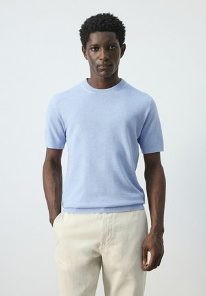 Mand med kort krøllet hår iført lyseblå kortærmet sweater og beige bukser, stående mod en ensartet lys grå baggrund.