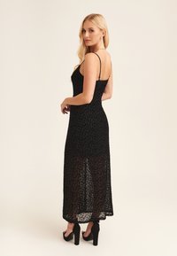 Black leopard print maxi dress with a fitted bodice, thin adjustable straps, and sheer lower hem. Paired with high block-heeled sandals.