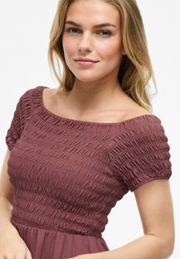 Robe bordeaux avec texture smockée, décolleté épaules nues, manches courtes bouffantes et jupe fluide. Tissu lisse avec un léger éclat.