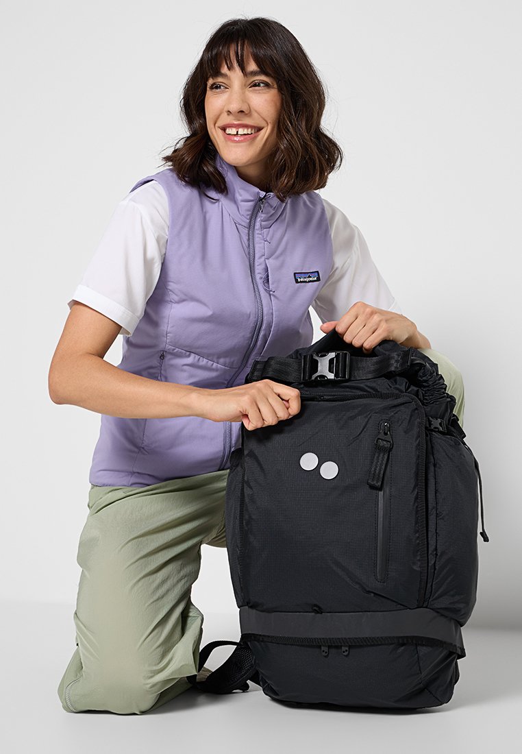Lächelnde Frau, die eine lavendelfarbene Weste und helle Hosen trägt, kniet und hält einen großen schwarzen Rucksack mit Schnallen und Reißverschlusstaschen geöffnet.