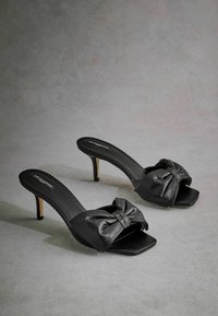 Mules à talons hauts en cuir noir avec un grand nœud sur le devant, bout carré et talon aiguille fin. Texture lisse avec un minimum de détails.
