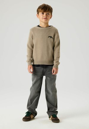Young boy standing wearing a beige sweater, black shirt underneath, gray jeans, and brown sneakers with green laces on white background.