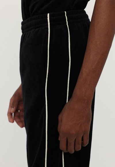 Jogging en velours noir avec des rayures latérales blanches, dotés d'une taille élastique et de poches, affichant une texture lisse et une coupe ajustée.