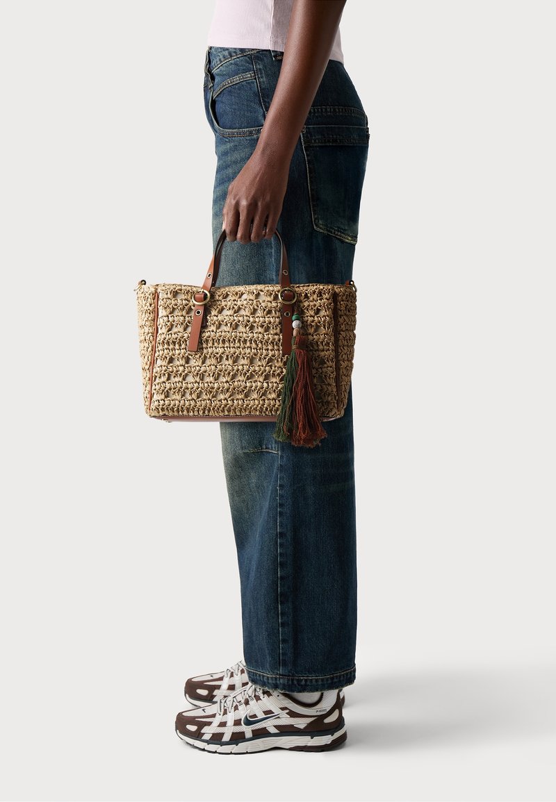 Personne portant un jean bleu et des baskets blanches tenant un sac à main en paille tressée avec des poignées en cuir et des pampilles décoratives.