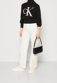 Maglione nero a coste con logo 'CK' bianco, abbinato a jeans bianchi a gamba larga e una piccola borsa nera con hardware argentato. Calzature casual.